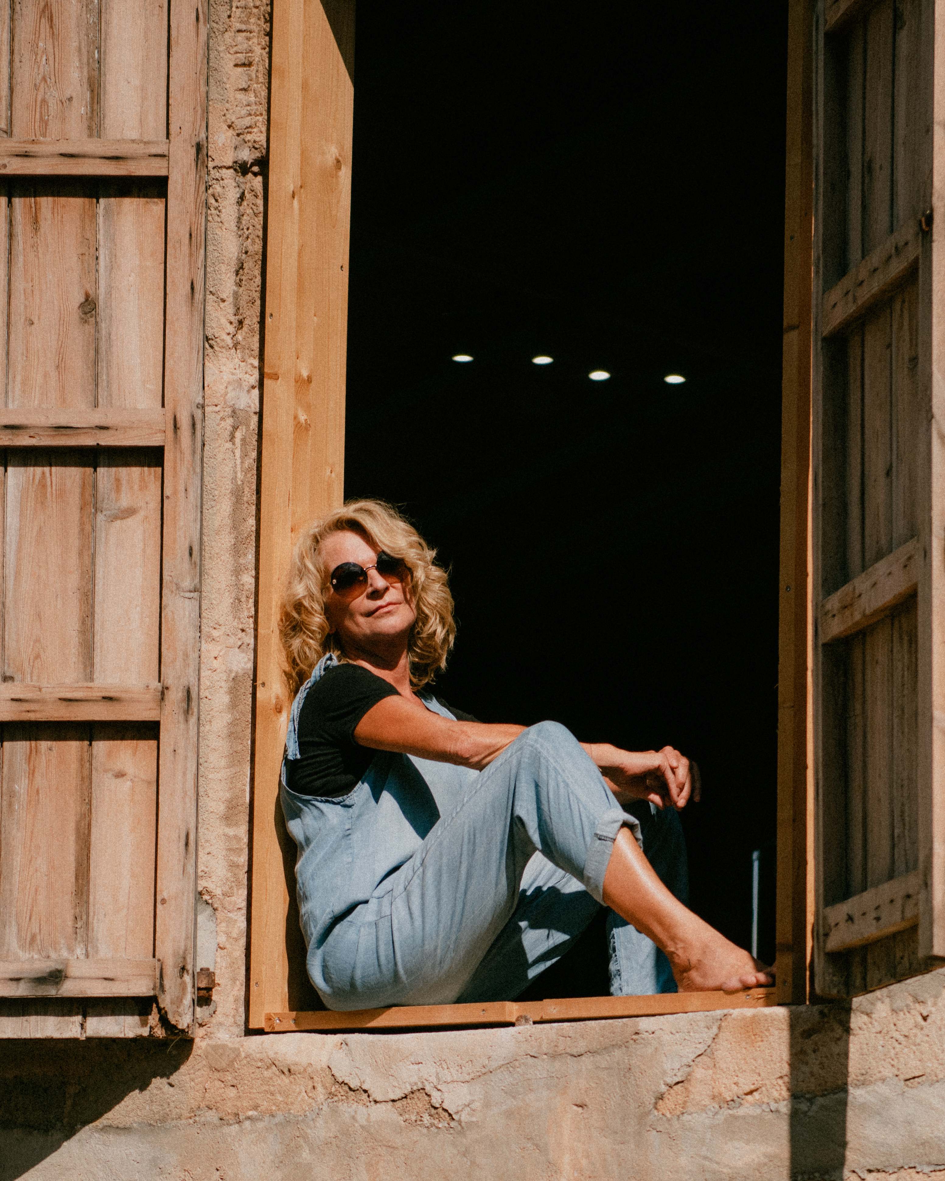 Karin Galler sitzt mit Sonnenbrille im Rahmen eines alten Holzfensters